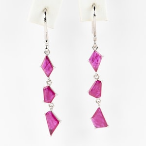 Könnte beinhalten: Ein Paar silberner Ohrhänger mit drei fuchsiafarbenen, geometrisch geformten Edelsteinen. Die Ohrringe hängen an einem silbernen Haken vor einem weißen Hintergrund.