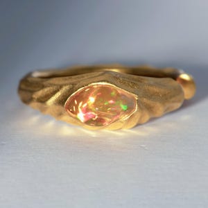 May include: A gold-colored ring with an oval-shaped opal gemstone. The opal displays a spectrum of colors, including red, green, and yellow. The ring has a textured band and a smooth, polished finish on the gemstone.