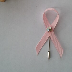 May include: A pink ribbon with a rhinestone in the center, attached to a silver pin. The ribbon is a symbol of breast cancer awareness.