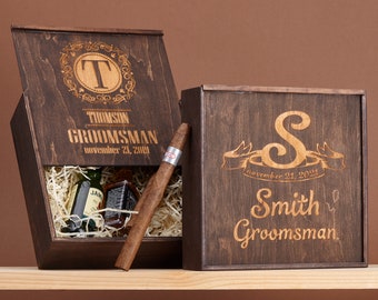 May include: Two wooden boxes with engraved personalised messages. The boxes are dark brown with a wood grain pattern. The top box is open and contains a cigar, two small bottles, and shredded paper. The bottom box is closed and has a ribbon design with the date "November 21, 2021".
