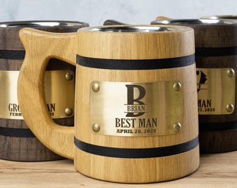 May include: A set of four wooden beer mugs with personalised engraved brass plates. The mugs are made of light brown wood with black bands around the top and bottom. The brass plates feature a letter, name, and the words "Best Man" and the date.