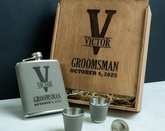 May include: A personalised groomsman gift set comprising a wooden box, a flask, and two shot glasses. The box and flask are engraved with the letter "V", the name "Victor", and the word "Groomsman" with a date. The flask and shot glasses have a grey leatherette wrap.