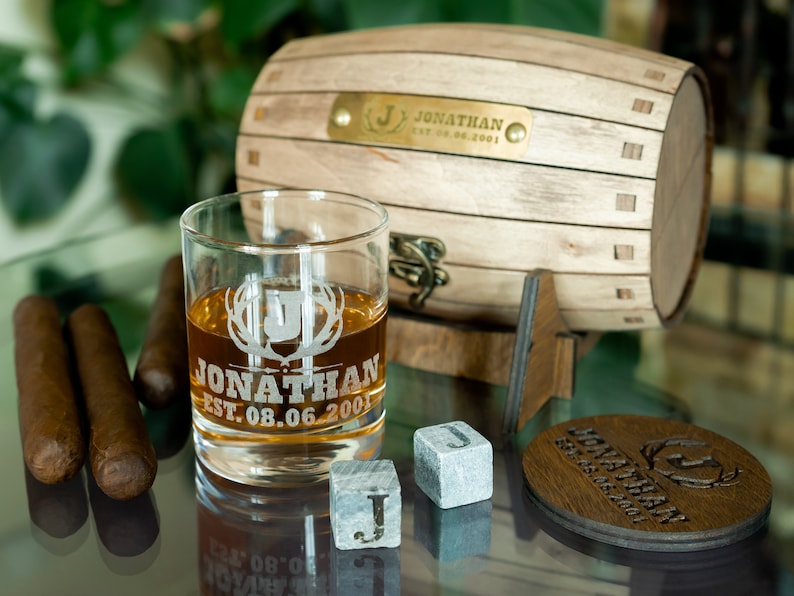 May include: A personalised whiskey glass with the name "Jonathan Est. 08.06.2001" etched on the side, along with a stag's head and antlers. The glass is filled with amber liquid and sits on a table with a wooden barrel, coasters, and two grey stone cubes.