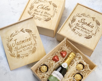 May include: Three wooden gift boxes with engraved floral wreaths and personalised names. The boxes are filled with champagne, chocolates, and straw. The text on the boxes reads "Chloe Maid of Honour June 12.2025", "Sophia Bridesmaid June 12.2025", and "Jasmine Bridesmaid June 12.2025".
