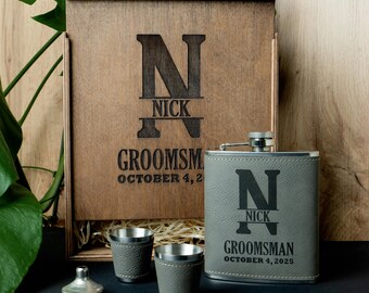 May include: A personalised groomsman gift set comprising a wooden box, a grey leather-wrapped flask, two shot glasses, and a funnel. The box and flask are engraved with the initial "N", the name "Nick", and the word "Groomsman" with a date.