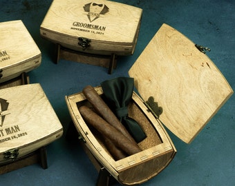 May include: Three wooden boxes with lids, each engraved with the word "Groomsman" and a date. One box is open and contains two cigars and a black bow tie.
