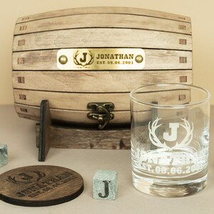 May include: Personalised whiskey gift set with a wooden barrel, a glass with antlers and a letter J, a coaster, and four whiskey stones. The barrel has a brass plaque with the text "Jonathan Est. 08.06.2001".