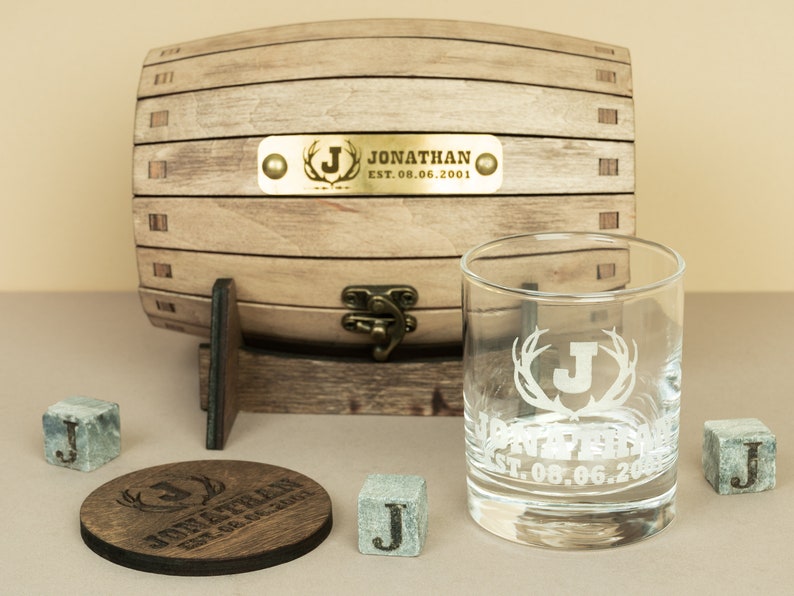 May include: Personalised whiskey gift set with a wooden barrel, a glass with antlers and a letter J, a coaster, and four whiskey stones. The barrel has a brass plaque with the text "Jonathan Est. 08.06.2001".