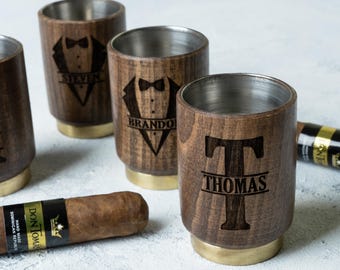 May include: Four wooden shot glasses with gold rims and personalised engravings. The glasses are arranged in a row on a white surface. The engraving on the glasses includes names and a tuxedo design. A brown cigar is also visible in the image.
