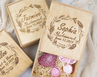 May include: Three wooden boxes with engraved floral wreaths and personalised text. The boxes are labelled "Jasmine Bridesmaid June 12, 2025", "Sophia Bridesmaid June 12, 2025", and "Chloe Maid of Honour June 12, 2025". The box in the centre is open and contains pink and white gifts.