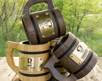 May include: Three wooden beer mugs with personalised engraved brass plates. The mugs are made of dark wood with black bands around the top and bottom. The plates have the words "Best Man" and "Groomsman" engraved on them, along with the names and dates.