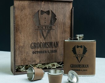 May include: A wooden gift box set with a hip flask, two shot glasses, and a funnel. The flask and shot glasses have brown leather detailing. The box and flask are engraved with a tuxedo design, the name "Victor", and the text "Groomsman October 4, 2025".