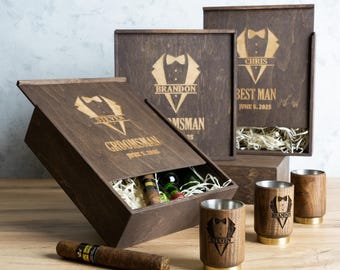 May include: Three wooden boxes with engraved tuxedo designs and names, 'Steven', 'Brandon', and 'Chris'. The box labelled 'Chris' also says 'Best Man' and 'June 5, 2025'. The boxes are open and contain a bottle of wine, a cigar, and a set of four wooden shot glasses.