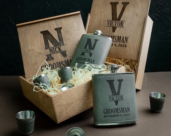 May include: Wooden gift boxes containing personalised flasks and shot glasses. The grey flasks and shot glasses have black stitching. The boxes are engraved with names, the word "Groomsman" and a date.