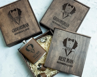 May include: Wooden gift boxes with engraved tuxedo designs and names, including "Victor," "Nick," and "Evan." The boxes contain a brown flask, shot glasses, and a small silver cup, all nestled in straw. The text "Groomsman" and "Best Man" are visible.
