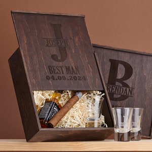 Puede incluir: Dos cajas de regalo de madera con iniciales y nombres grabados. La caja de la izquierda tiene una botella de whisky, un cigarro y un vaso de chupito dentro. La caja de la derecha tiene dos vasos de chupito dentro. Las cajas son marrones y tienen una textura de madera.