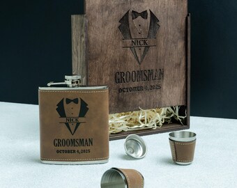 May include: A brown leather-covered hip flask and shot glasses set, presented with a wooden box. The flask and box are decorated with a tuxedo design and the name "NICK" with "GROOMSMAN OCTOBER 4, 2025" engraved. The box is open, revealing straw.