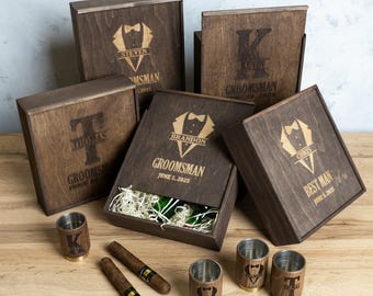 May include: Wooden gift boxes with personalised engravings for a groomsman, best man, and other wedding party members. Each box includes a shot glass and a cigar. The boxes are decorated with a tuxedo graphic and the names of the recipients.