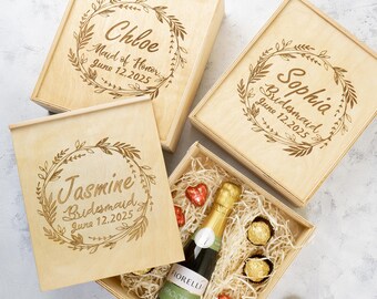 May include: Four wooden gift boxes with engraved floral wreaths and personalised text. The boxes are labelled "Chloe Maid of Honour June 12, 2025", "Sophia Bridesmaid June 12, 2025", and "Jasmine Bridesmaid June 12, 2025". One box is open and contains a bottle of sparkling wine, chocolate truffles, and shredded paper.