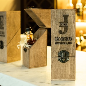 Puede incluir: Cajas de regalo de madera con grabado personalizado. Una caja está abierta, revelando una pequeña botella de licor metida en virutas de madera. La parte frontal de la caja dice "Josh GROOMSMAN November 16, 2024".