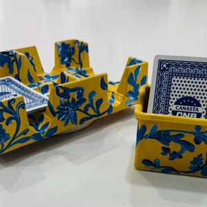 May include: Yellow and blue painted card holders with a floral design. One holder is rectangular with multiple compartments, holding playing cards. The other is a smaller rectangular holder with a deck of cards. The cards have a blue and white design.