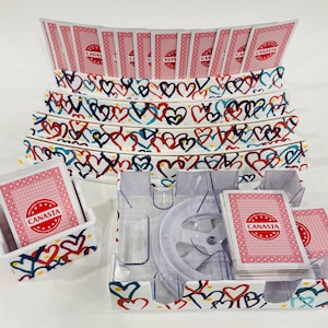 May include: A Canasta card game set with a clear plastic card holder and several decks of cards. The card holders and card trays are decorated with a pattern of colorful hearts. The cards have the word "CANASTA" printed on them.
