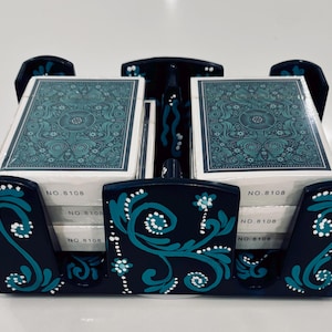 May include: A dark blue wooden playing card holder featuring hand-painted teal and white floral scrollwork designs. The holder contains two stacks of playing cards, each wrapped in clear plastic with the text NO. 8108 printed on the side.