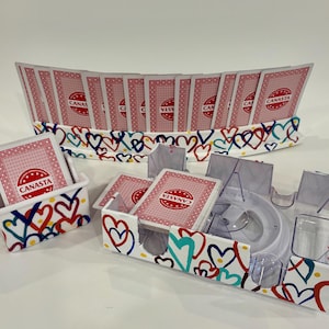 May include: A Canasta card game set with a decorative heart design. The set includes card holders, a card tray, and a card rack, all adorned with colorful hearts. The cards are red with the word "CANASTA" printed on them.