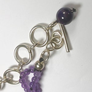May include: A silver chain bracelet with a purple amethyst bead toggle clasp. The bracelet is made of small, round amethyst beads and silver chain links.