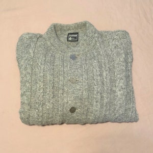 Könnte beinhalten: Ein grauer Strickpullover mit vier silbernen Knöpfen. Der Pullover hat einen gerippten Kragen und Bündchen.