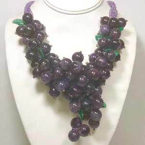 May include: A purple amethyst grape necklace with green leaves. The necklace is made of faceted amethyst beads and is strung on a silver chain.