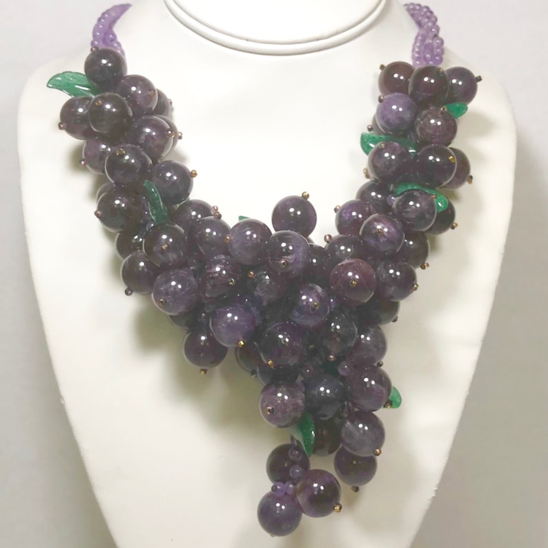 May include: A purple amethyst grape necklace with green leaves. The necklace is made of faceted amethyst beads and is strung on a silver chain.