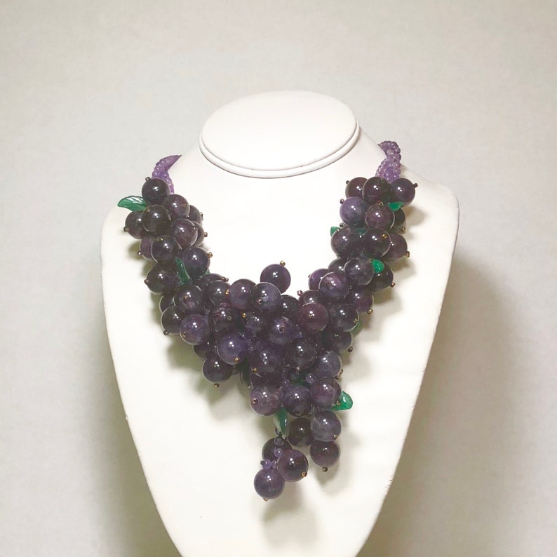 May include: A purple amethyst grape necklace with green leaves. The necklace is made of faceted amethyst beads and is strung on a silver chain.