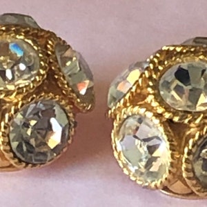 Pode incluir: Um par de brincos de pressão dourados. Cada brinco apresenta um aglomerado de cristais transparentes e facetados, inseridos em uma borda texturizada em forma de corda. Os cristais refletem a luz, criando um efeito cintilante.