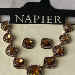 Puede incluir: Un collar dorado y un juego de pendientes cuadrados a juego. La joyería presenta piedras facetadas de color ámbar engastadas en un marco cuadrado con un borde en forma de cuerda. La marca "NAPIER" está impresa en el embalaje.