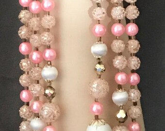 May include: A vintage necklace with three strands of beads. The necklace features pink, clear, and white beads, with gold-colored accents. The beads are arranged in a layered design, creating a classic and elegant look. The necklace is displayed on a mannequin.