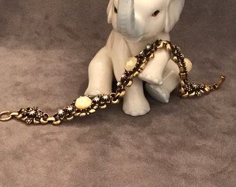 Puede incluir: Una figura de elefante de cerámica blanca con la trompa levantada sostiene una pulsera dorada. La pulsera presenta detalles florales y de piedras preciosas. El elefante tiene ojos rojos y una trompa gris.