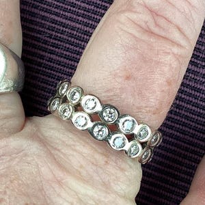 May include: A silver ring with two rows of clear, round gemstones. The ring is worn on a finger, with another silver ring visible on an adjacent finger. The background is a textured, dark purple fabric.