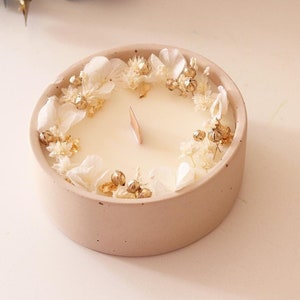 May include: A round, light pink candle in a matching holder, decorated with white flowers and gold accents. The candle has a wooden wick and a creamy white wax. The word "AYANA" is printed at the bottom left.