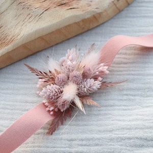 Könnte beinhalten: Ein rosa Band mit einem kleinen Strauß getrockneter Blumen in Rosa- und Weißtönen. Der Strauß enthält getrocknete Gräser und flauschige Blumen.