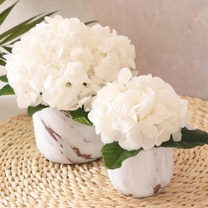 Peut inclure: Deux pots en céramique blanche avec un design marbré, chacun contenant un bouquet d'hortensias blancs. Les pots sont posés sur un tapis de paille tressé.
