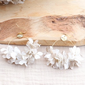 May include: A pair of gold hoop earrings with white dried hydrangea flowers attached. The earrings are on a wooden surface.
