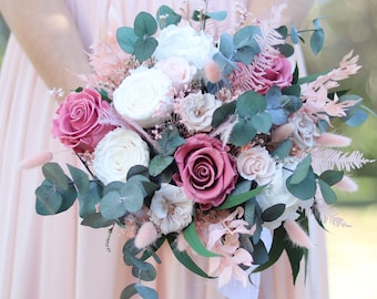 May include: A bouquet of preserved roses in shades of pink and white, with eucalyptus greenery and dried flowers. The bouquet is tied with a white ribbon and has the word "AYANA" written on the bottom.