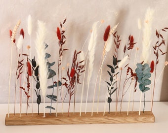 Può includere: Un supporto in legno con una varietà di fiori e erbe secche, tra cui erba della pampa bianca, fiori secchi rossi e marroni e foglie di eucalipto verdi.