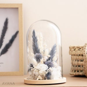 May include: A decorative floral arrangement encased in a glass dome. The arrangement features dried flowers in shades of white, blue, and beige, set on a wooden base. A framed print and a woven basket are in the background.
