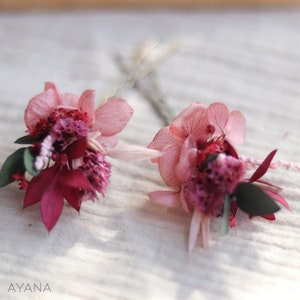 May include: Two pink and burgundy dried flower hair clips. The flowers are arranged in a cluster and are attached to a silver hairpin.