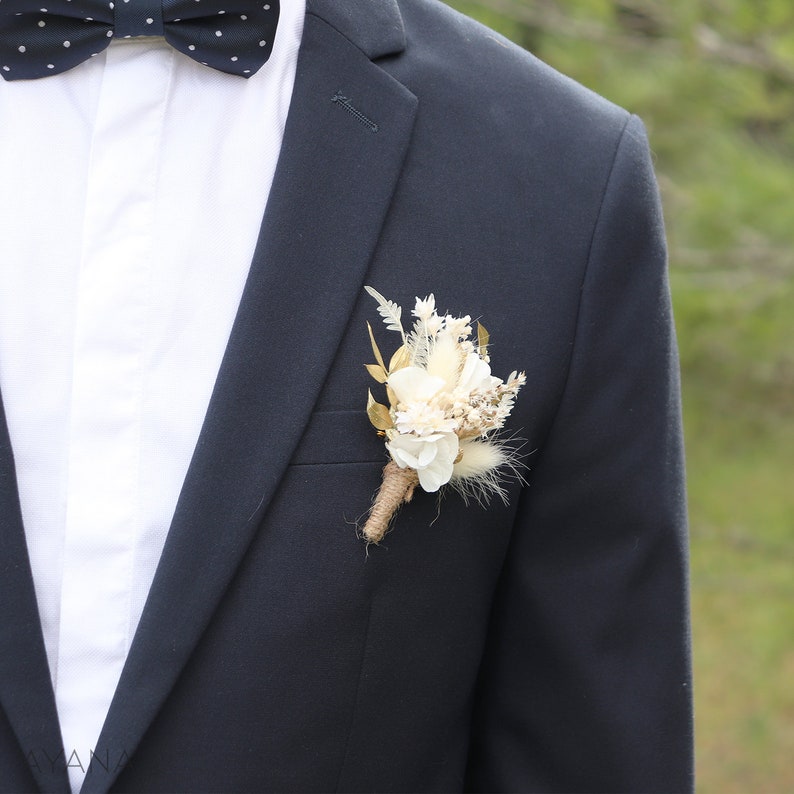 May include: A white and beige dried flower buttonhole with gold accents, ideal for a wedding or special occasion.