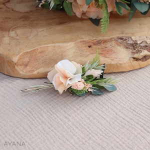 May include: A small bouquet of peach and white silk flowers with green leaves and stems. The bouquet is attached to a gold hairpin.