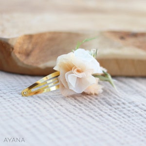 May include: A delicate hair clip featuring a cluster of beige and white fabric flowers. The clip is gold-toned and has a simple design.