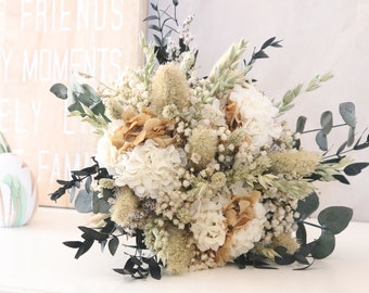 Puede incluir: Un ramo de flores secas con hortensias blancas, proteas color beige y hojas de eucalipto verdes. El arreglo incluye pequeñas flores de relleno blancas y acentos verde oscuro, creando un aspecto natural y texturizado. El ramo se exhibe sobre una superficie blanca.